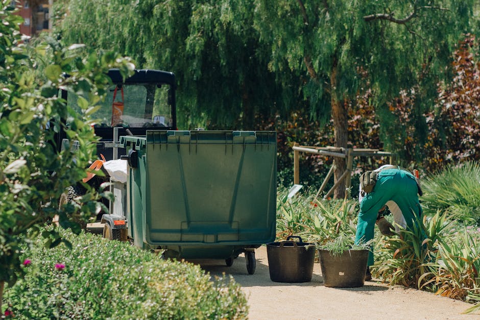 A gardener wearing green overalls is engaged in hedge trimming in a lush, well-maintained garden. The scene features neatly arranged garden beds with green, spiky foliage, along with a variety of plan